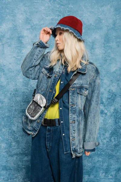 Blonde woman in denim jacket adjusting trendy panama hat near blue textured background — Stock Photo