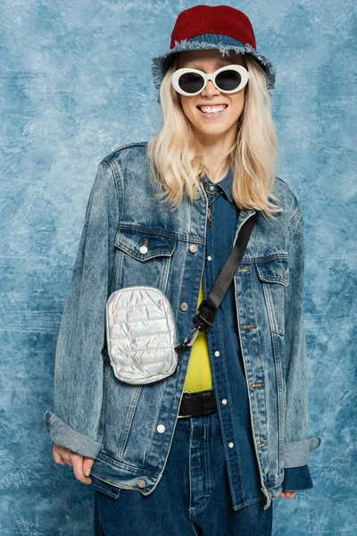 Счастливая блондинка в джинсовой одежде позирует в панамской шляпе и солнечных очках рядом с синим текстурированным фоном — стоковое фото