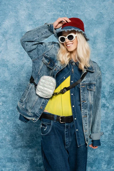 Cheerful blonde woman in denim outfit posing in panama hat and sunglasses near blue textured background — Stock Photo