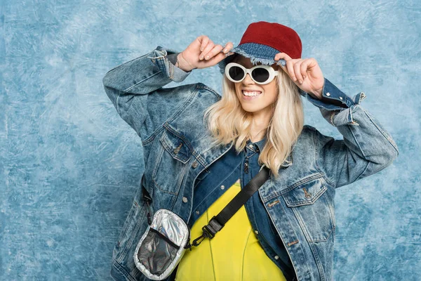 Smiling blonde woman in denim jacket and sunglasses adjusting panama hat near blue textured background — Stock Photo