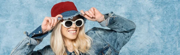 Smiling blonde woman in denim jacket and sunglasses adjusting panama hat near blue textured background, banner — Stock Photo