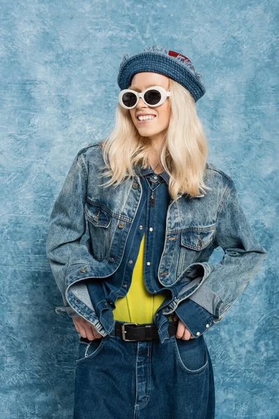 Счастливая блондинка в джинсовой шляпе Панама и солнцезащитных очках позирует рядом с синим текстурированным фоном — стоковое фото