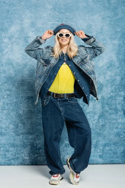 Full length of happy blonde woman in denim panama hat and sunglasses posing near blue textured background — Stock Photo