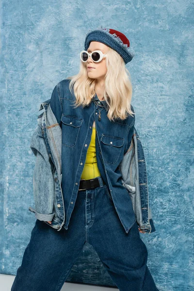 Блондинка в джинсовой шляпе и солнцезащитных очках на фоне синего текстурированного фона — стоковое фото