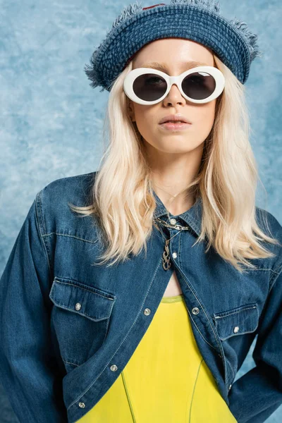 Young blonde woman in denim panama hat and sunglasses posing near blue textured background — Stock Photo
