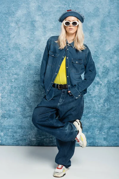 Full length of blonde woman in denim outfit posing in panama hat and sunglasses near blue textured background — Stockfoto