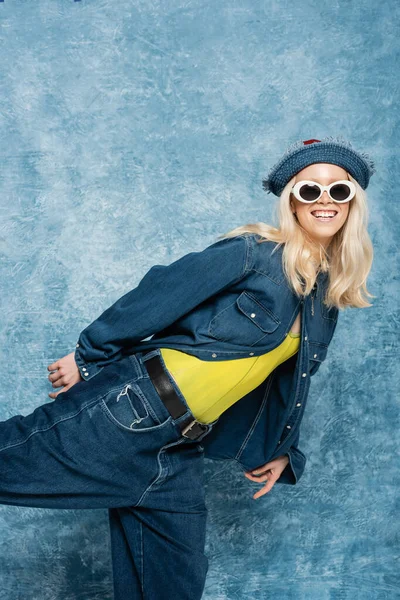 Довольная блондинка в джинсовой шляпе и солнцезащитных очках позирует рядом с синим текстурированным фоном — стоковое фото