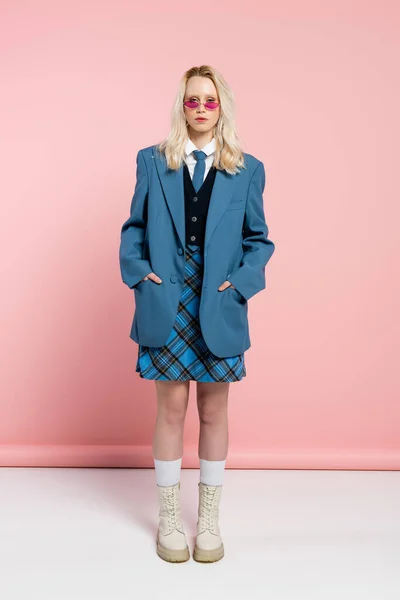 Full length of blonde woman in blue blazer and plaid skirt standing with hands in pockets on pink — Stock Photo