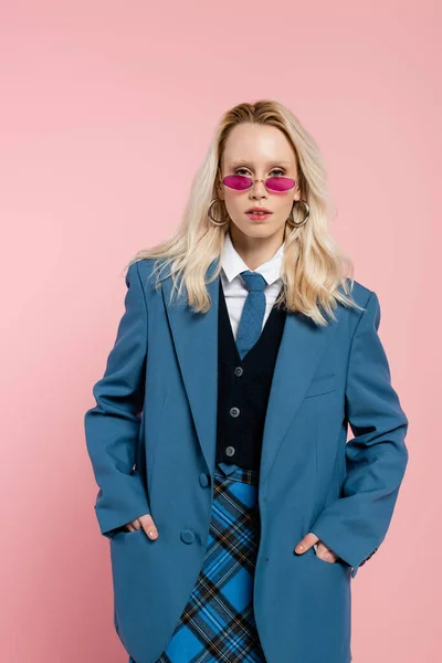 Blonde woman in blue blazer with tie and sunglasses posing with hands in pockets isolated on pink — Stock Photo