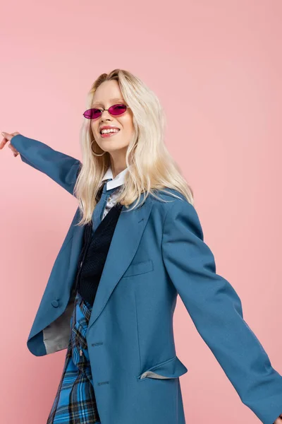 Lächelnde Frau im blauen Blazer mit Krawatte und Sonnenbrille posiert mit ausgestreckter Hand isoliert auf rosa — Stockfoto