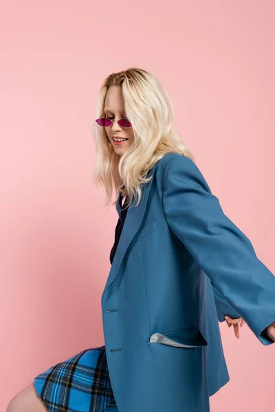 Mulher loira em blazer azul e óculos de sol elegantes posando isolado em rosa — Fotografia de Stock