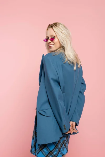 Femme blonde heureuse en tenue bleue et lunettes de soleil tendance posant isolée sur rose — Photo de stock