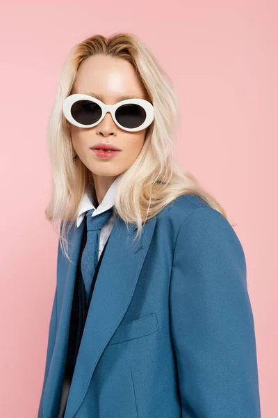 Young blonde woman in blue blazer and sunglasses looking at camera isolated on pink — Stock Photo