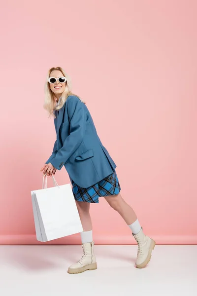 In voller Länge glückliche Frau im blauen Outfit mit kariertem Rock und trendiger Sonnenbrille posiert mit Einkaufstaschen auf rosa — Stockfoto