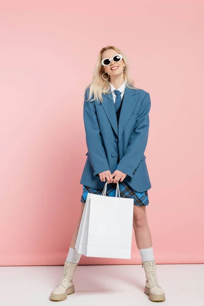 Volle Länge der fröhlichen Frau in blauem Outfit mit kariertem Rock und trendiger Sonnenbrille posiert mit Einkaufstaschen auf rosa — Stockfoto