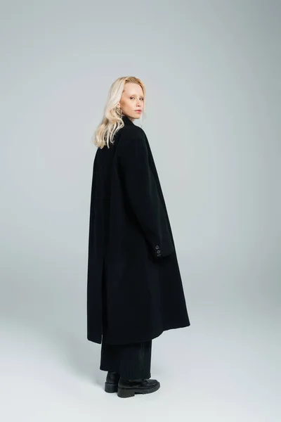 Full length of blonde young woman in black coat standing on grey — Fotografia de Stock