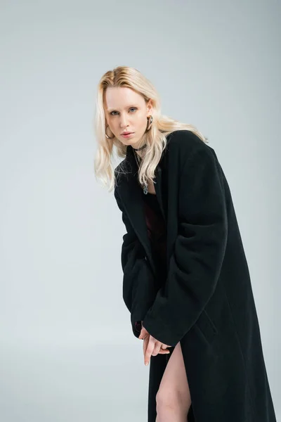 Blonde woman in black skirt and coat posing while looking at camera isolated on grey — Stockfoto