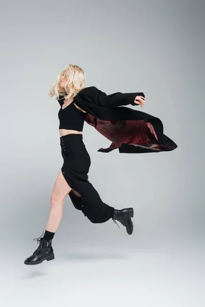Comprimento total de loira e jovem mulher na roupa preta elegante pulando em cinza — Fotografia de Stock
