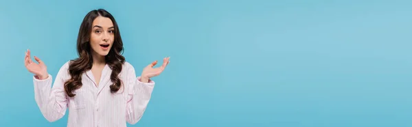 Surprised young woman in sleepwear gesturing while standing with opened mouth isolated on blue, banner — Stock Photo