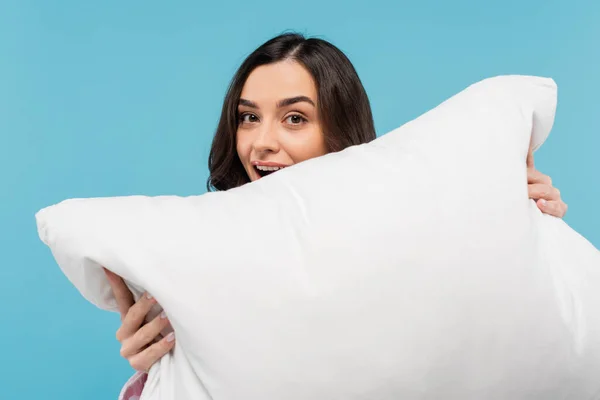 Здивована молода жінка з відкритим ротом тримає білу подушку ізольовано на синьому — стокове фото