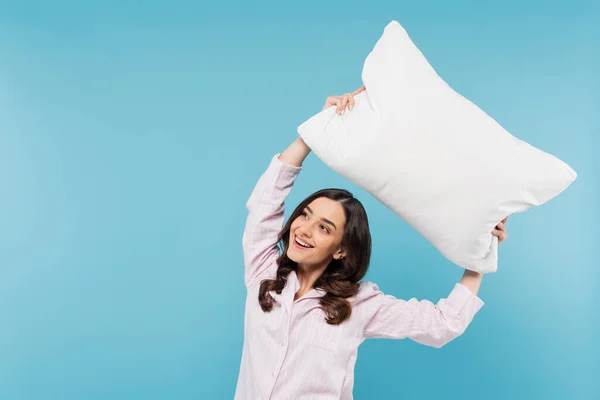 Весела молода жінка в сплячому одязі тримає білу подушку над головою ізольовано на синьому — стокове фото