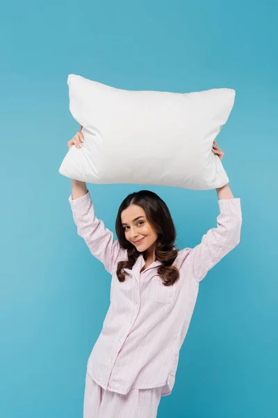 Щаслива молода жінка в сплячому одязі тримає білу подушку над головою ізольовано на синьому — стокове фото