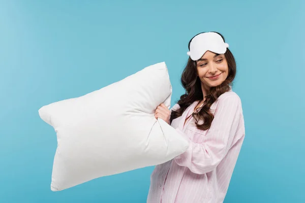 Задоволена молода жінка в піжамі і нічній масці, що тримає білу подушку ізольовано на синьому — стокове фото