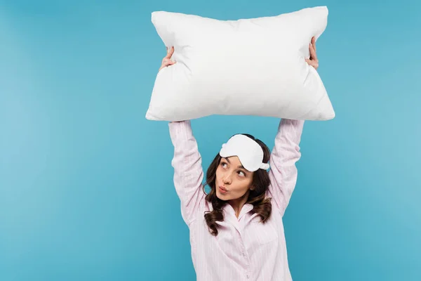 Brunette young woman in pajamas and night mask holding white pillow while pouting lips isolated on blue - foto de stock