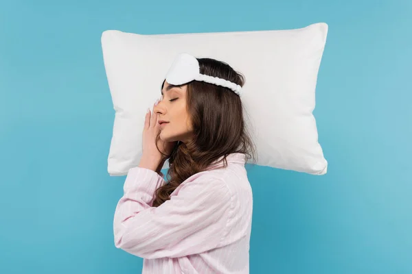 Vista lateral de mujer morena en pijama y máscara de noche durmiendo en almohada blanca aislada en azul - foto de stock