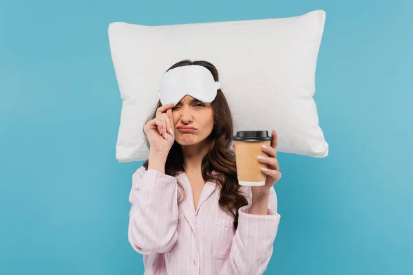 Femme mécontente en pyjama et masque de sommeil tenant tasse en papier avec café pour aller près oreiller volant isolé sur bleu — Photo de stock