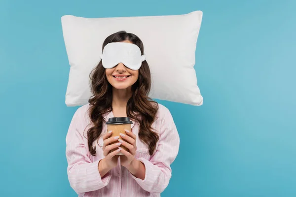 Pleased woman in pajamas and night mask holding paper cup with coffee to go near white pillow isolated on blue — Stock Photo