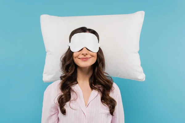 Pleased young woman in pajamas and night mask resting on white pillow isolated on blue — Stockfoto