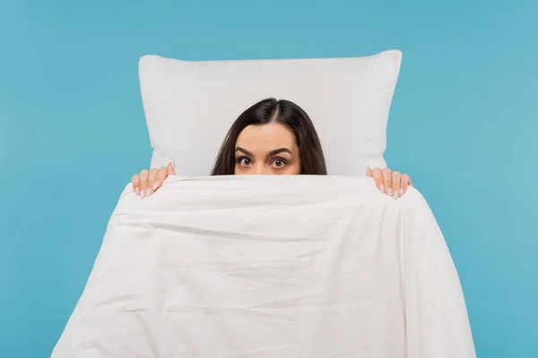 Young woman in pajamas covering face with warm duvet near pillow isolated on blue — Photo de stock