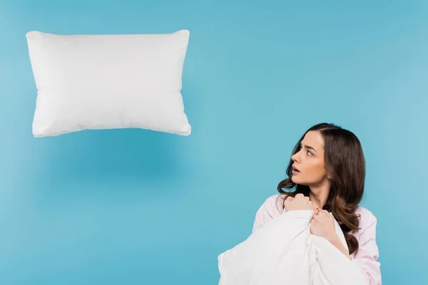 Mujer conmocionada en pijama sosteniendo el edredón caliente mirando la almohada levitante en azul - foto de stock