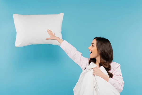 Mujer excitada en pijama sosteniendo un edredón caliente cerca de la almohada voladora en azul - foto de stock