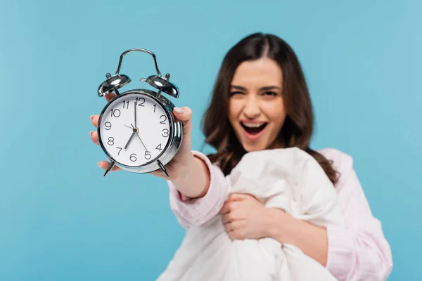 Amazed woman in pajamas holding vintage alarm clock and warm duvet isolated on blue — Foto stock