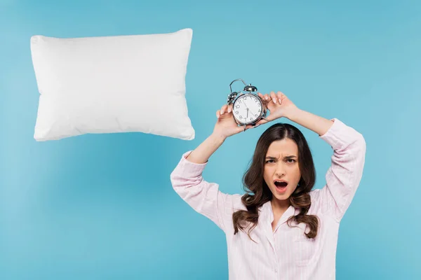 Shocked woman in pajamas holding vintage alarm clock above head near levitating pillow on blue — Stock Photo