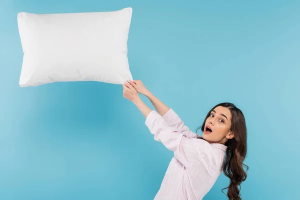 Shocked woman in pajamas pulling flying pillow on blue background — Photo de stock