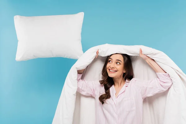 Cheerful woman in pajamas covering head with duvet and looking at flying pillow on blue background - foto de stock