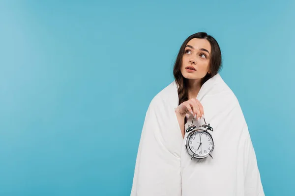 Dreamy young woman covered in white duvet holding vintage alarm clock isolated on blue — Stock Photo