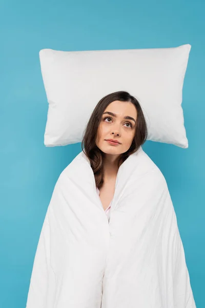 Dreamy young woman covered in white duvet standing near flying white pillow on blue background — Photo de stock