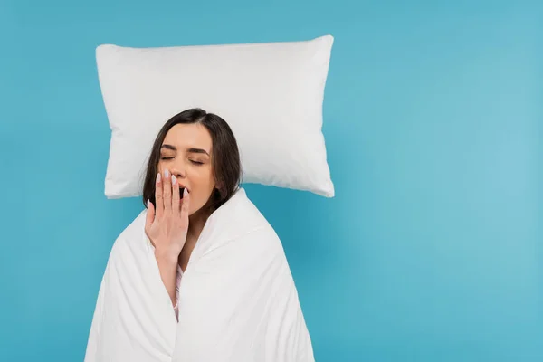 Tired woman covered in white duvet standing near flying white pillow and yawning on blue background — Stock Photo