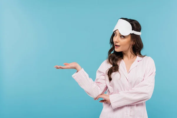 Brunette young woman in night mask and pajamas looking away and pointing with hand isolated on blue — Stock Photo