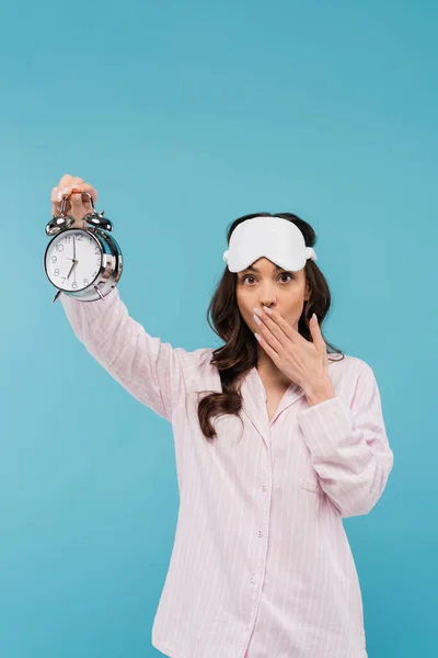 Shocked young woman in night mask and sleepwear holding alarm clock and covering mouth isolated on blue — Stock Photo