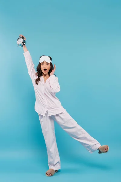 Volle Länge der schockierten jungen Frau in Nachtmaske und Schlafanzügen, die den Wecker auf blau hält — Stockfoto