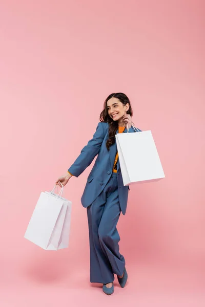 Full length of young and cheerful woman in blue pantsuit holding shopping bags on pink — Stock Photo