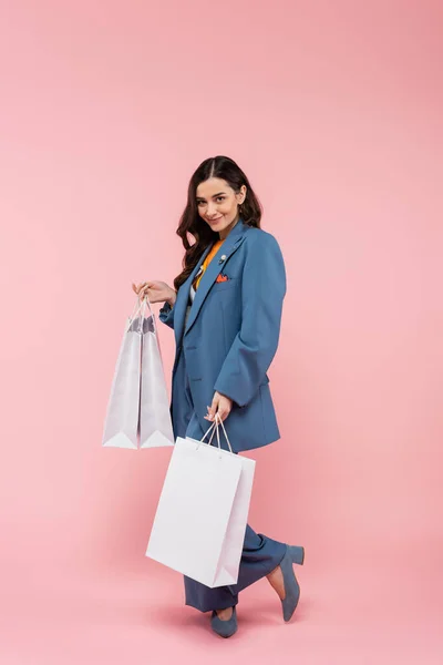 Full length of young and smiling woman in blue pantsuit holding shopping bags on pink — Stockfoto