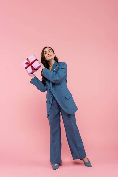 Longitud completa de la mujer complacida en traje de pantalón azul con regalo envuelto con cinta en rosa - foto de stock