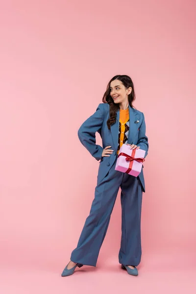 Full length of joyful woman in blue pantsuit holding wrapped present and standing with hand on hip on pink — Stock Photo