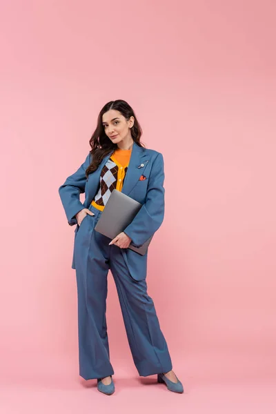 Full length of young brunette freelancer in blue pantsuit holding laptop while posing with hand in pocket on pink — Stock Photo
